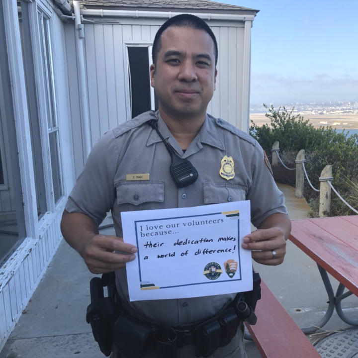 An up-close man with a Park Ranger uniform smiles outside in between an office building and a red picnic table and holds up a sign that says, “I love our volunteers because…their dedication makes a world of difference!”