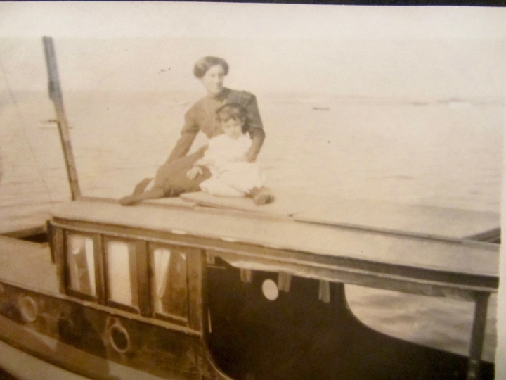 Celia Sweet wearing a dark dress standing behind her daughter Verla in a white dress sitting on top of a roof aboard her boat, Relue with water behind them.