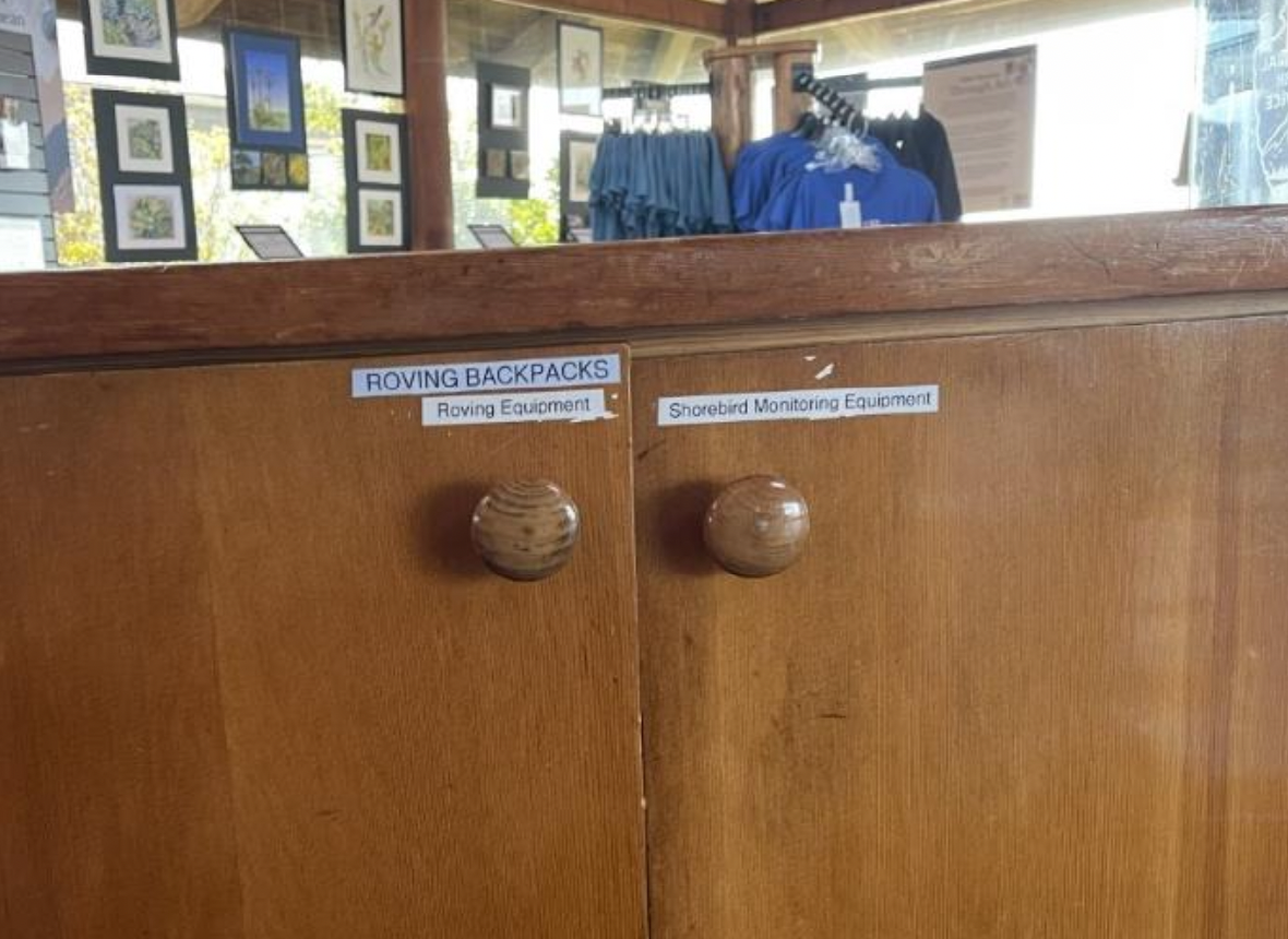 (Left) Cabinet behind the Visitor Center Information Desk with closed doors labeled with, “Roving Backpacks,” “Roving Equipment,” and “Shorebird Monitoring Equipment.”